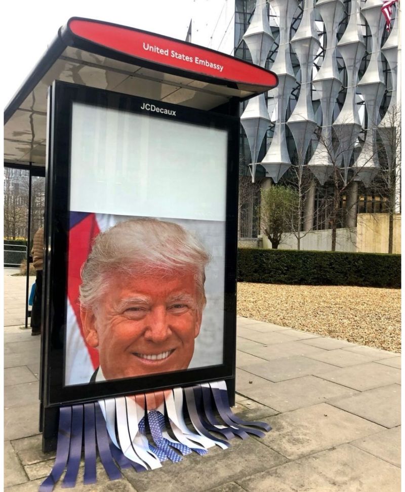 The image shows a bus shelter with a digital display board featuring a large portrait image of former U.S. President Donald Trump smiling and wearing a suit and tie being shredded to the floor. The background shows the distinctive architecture of the U.S. Embassy building in London.